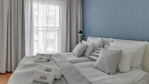 Ein Hotelzimmer mit einem großen Bett, weißer Bettwäsche und Blick auf die Stadt durch transparente Vorhänge.