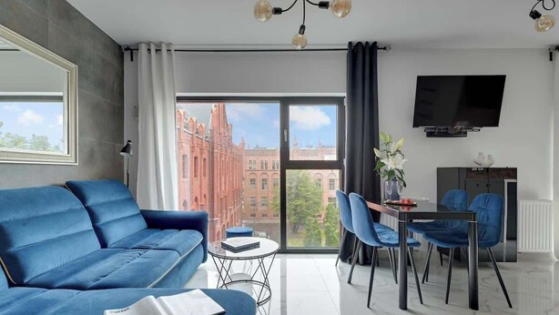 A modern living room with a blue sofa, a dining table with chairs, a flat-screen TV, and a large window with a city view. - Rezydencja Wintera 53 | Riverside (Gdańsk)