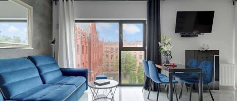 A modern living room with a blue sofa, a dining table with chairs, a flat-screen TV, and a large window with a city view.