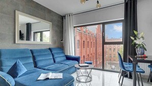 A modern living room with a blue sofa, a glass coffee table, and a dining area with a table and chairs. - Rezydencja Wintera 53 | Riverside (Gdańsk)