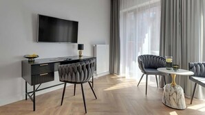 Een moderne hotelkamer met een flatscreen televisie, een zwart bureau met laden, twee gestoffeerde fauteuils, een marmeren salontafel en een groot raam met gordijnen.
