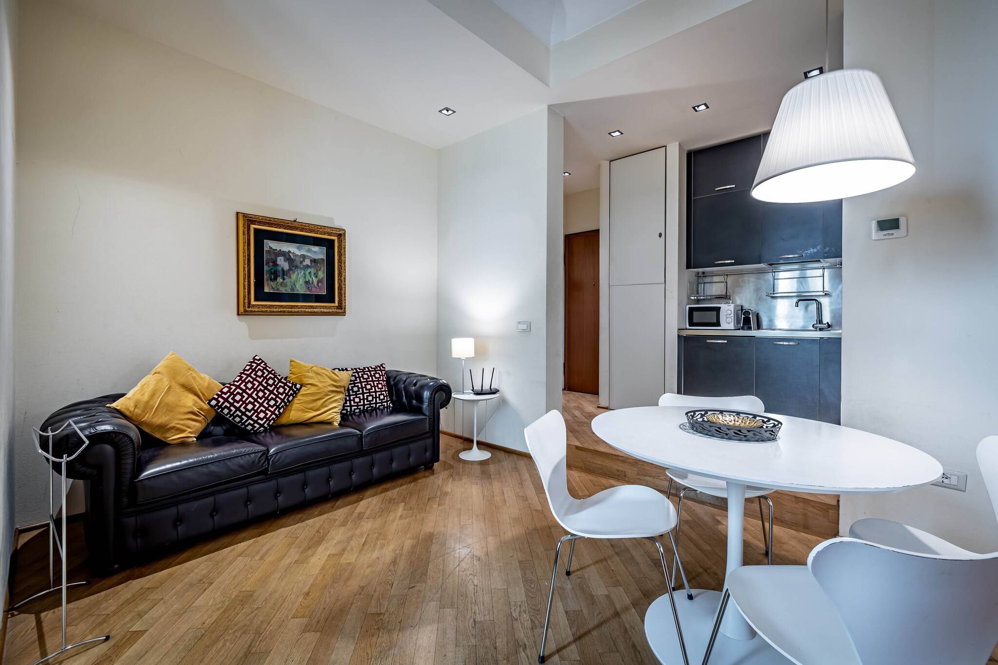 A modern living room with a black leather sofa, white chairs, a round table, and a kitchen area with dark cabinets and a microwave.
