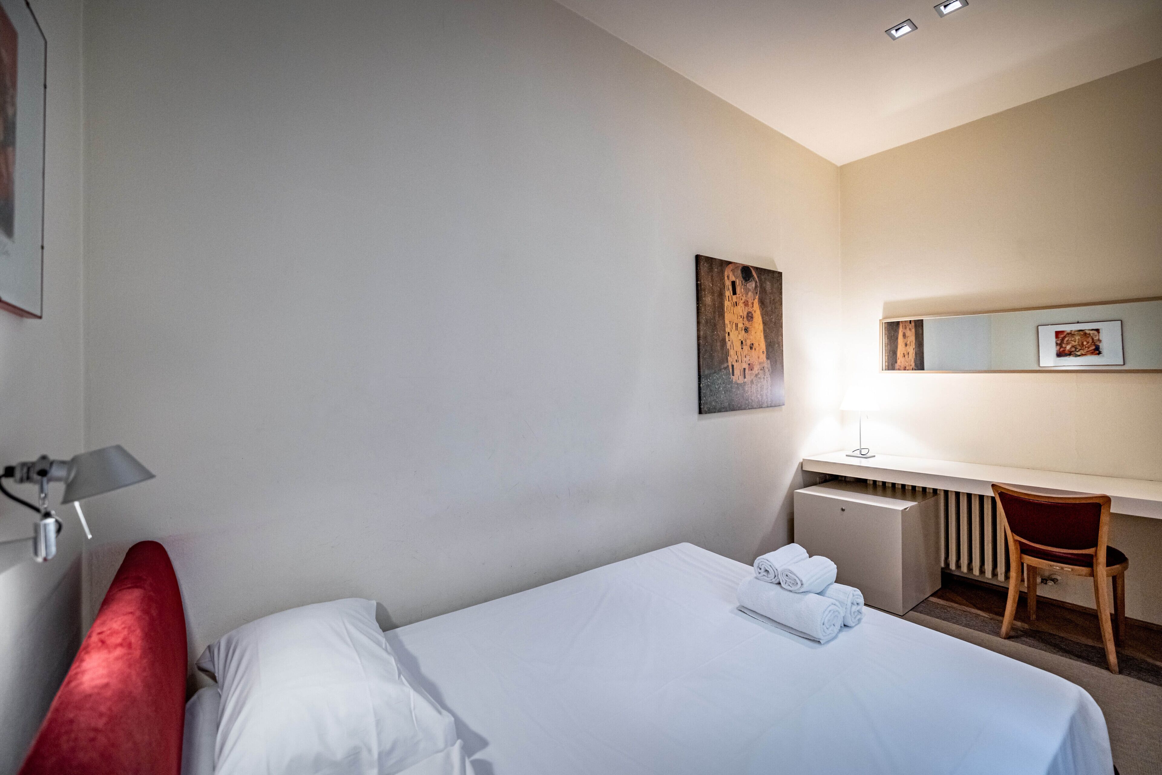 A hotel room with a bed, a desk, a chair, a radiator, and a painting on the wall.