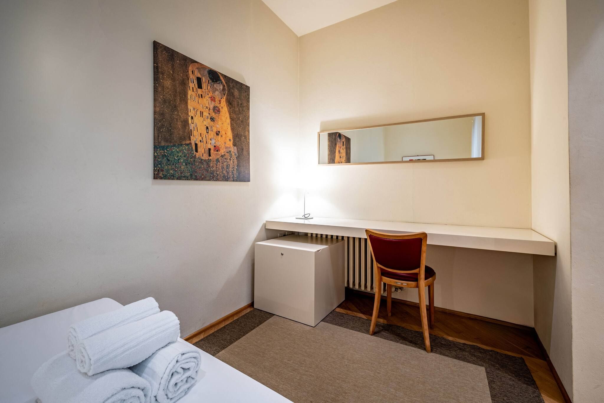 Een hotelkamer met een bed, een bureau met stoel, een radiator en een schilderij aan de muur.