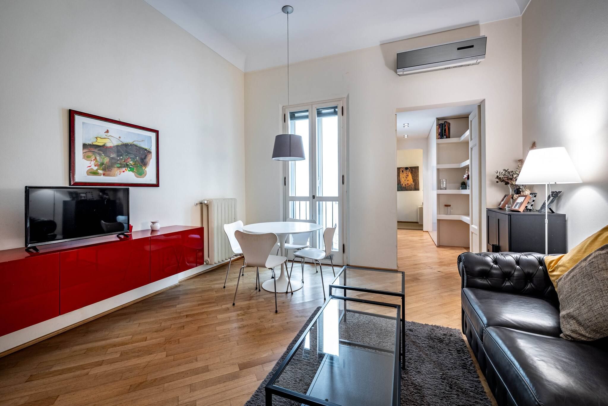 A modern living room with a red cabinet, a glass coffee table, a black leather sofa, and a dining area with white chairs.