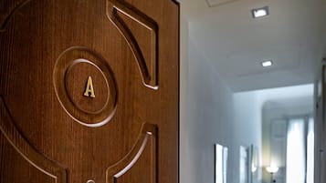 Una puerta de madera con un emblema circular y una cerradura, que da a un pasillo con suelo de madera y una pequeña mesa con una lámpara.