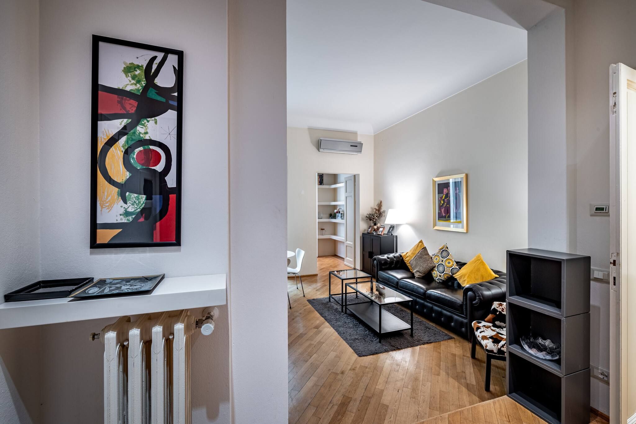 A modern living room with a black leather sofa, a coffee table, and a black rug. There is a white shelf with a framed picture and a radiator below it.