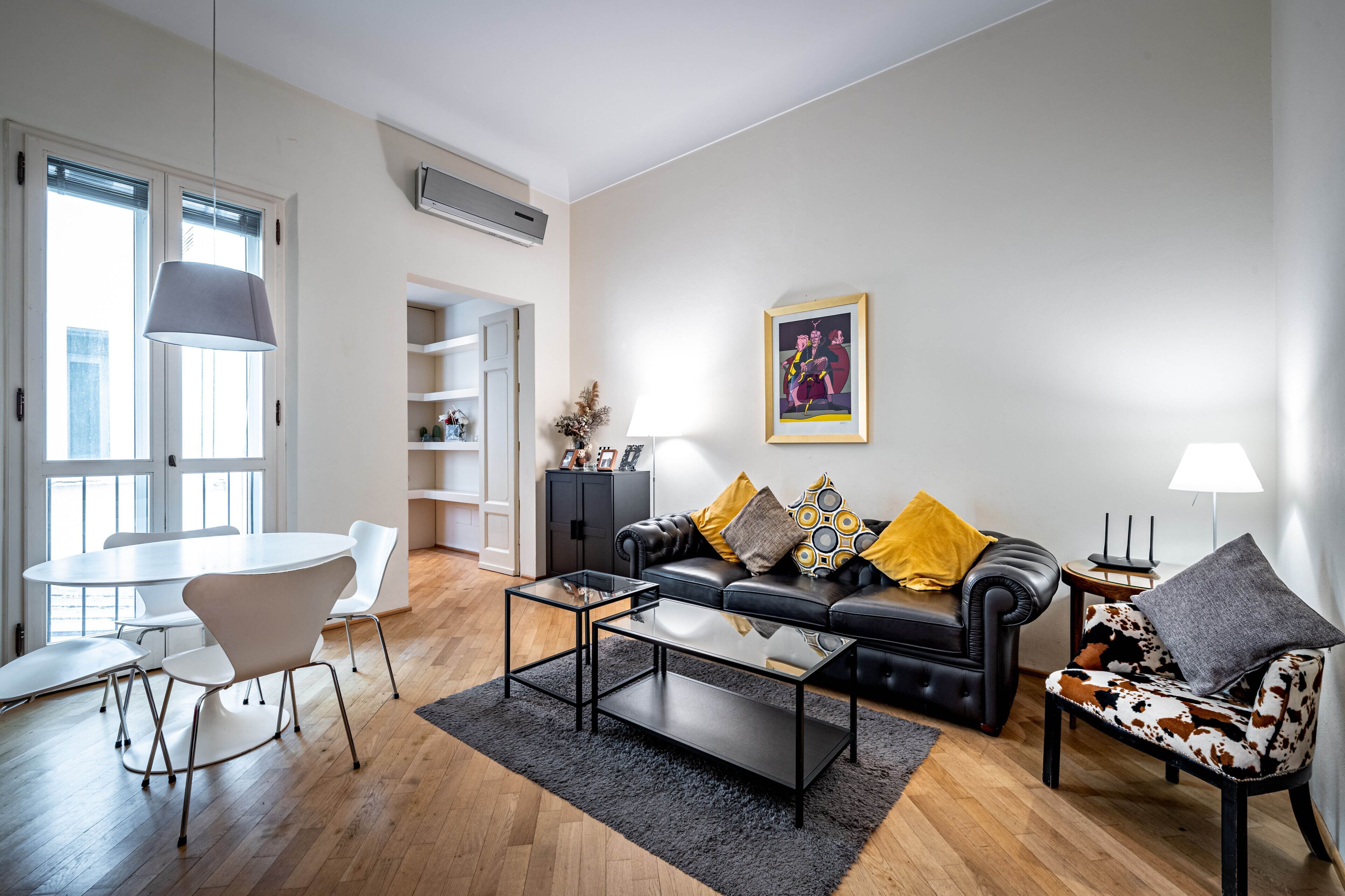A modern living room with a black leather sofa, a glass coffee table, a white dining table with chairs, and a grey rug.