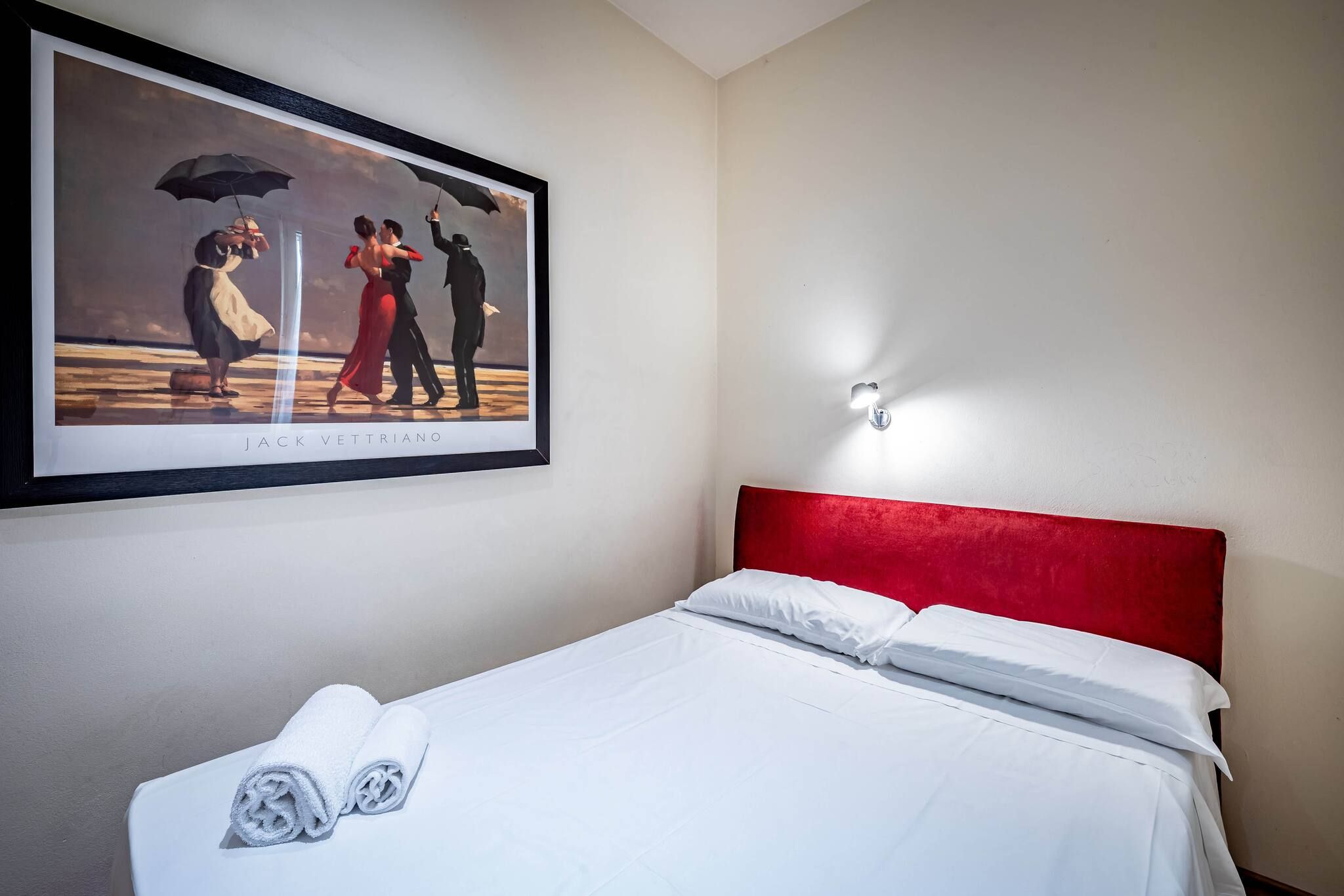 A hotel room with a bed, white linens, a red headboard, and a framed picture on the wall.
