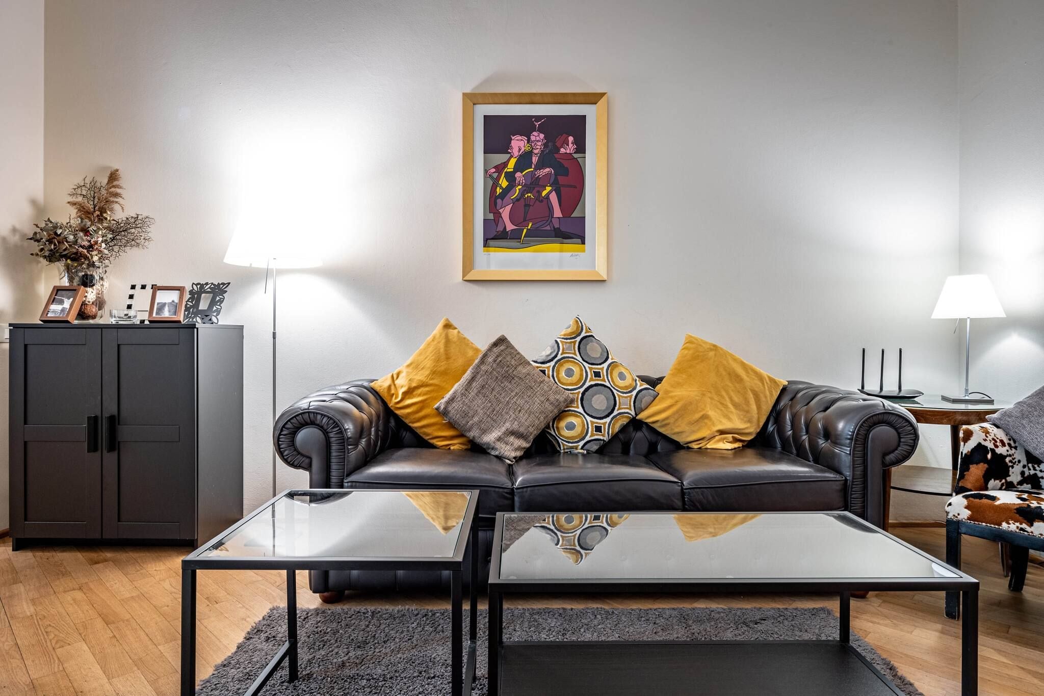 Ein modernes Wohnzimmer mit einem schwarzen Ledersofa, zwei Glastischen und einem gerahmten Kunstwerk an der Wand.