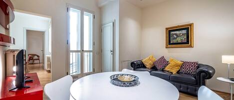 A modern living room with a round white table, a black leather sofa with patterned cushions, a framed picture on the wall, and a red accent wall.