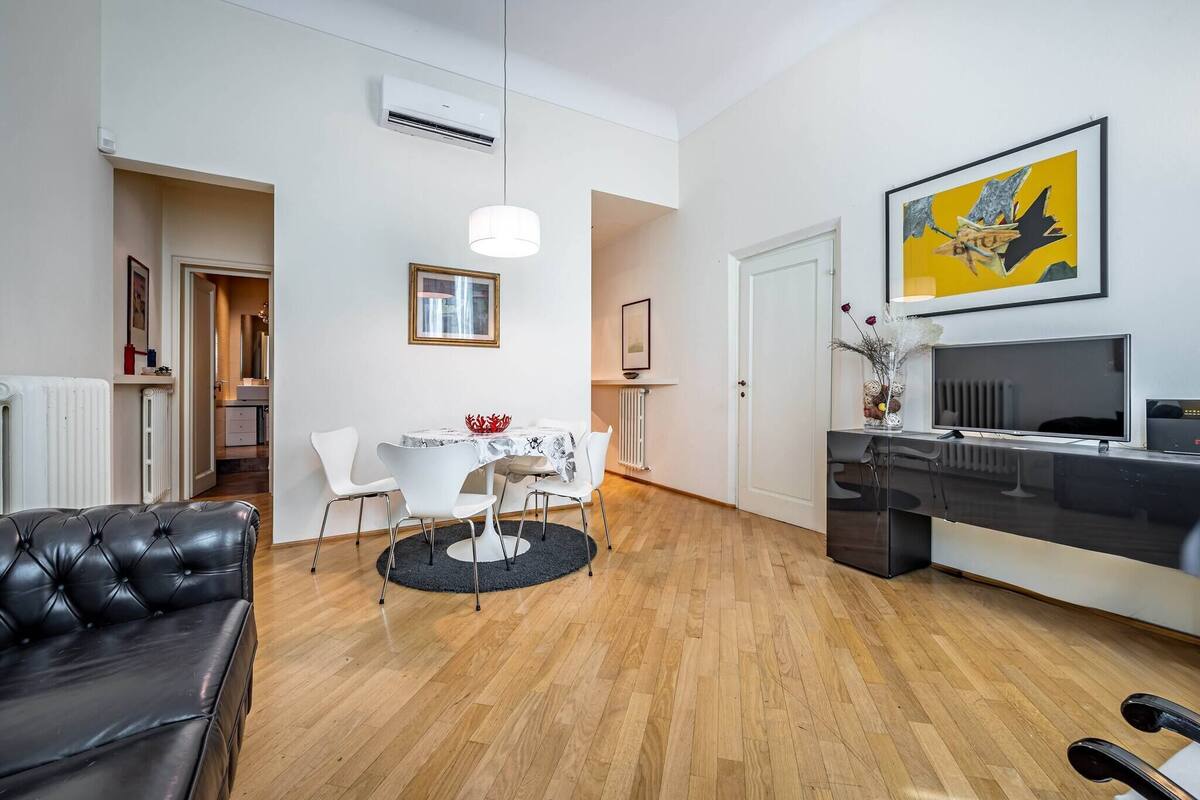 Un salon moderne avec un canapé en cuir noir, une table à manger avec des chaises blanches, une console noire et blanche, et une œuvre d’art encadrée accrochée au mur.