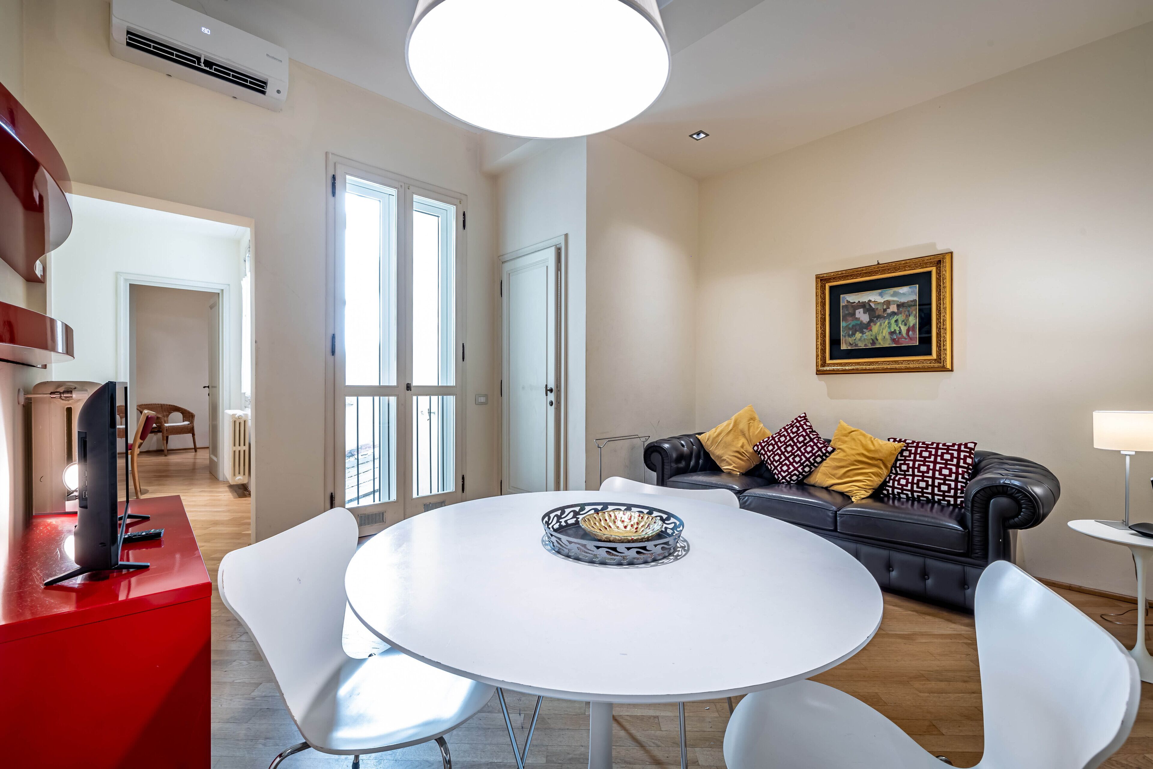 A modern living room with a round white table, a black leather sofa with patterned cushions, and a framed picture on the wall.