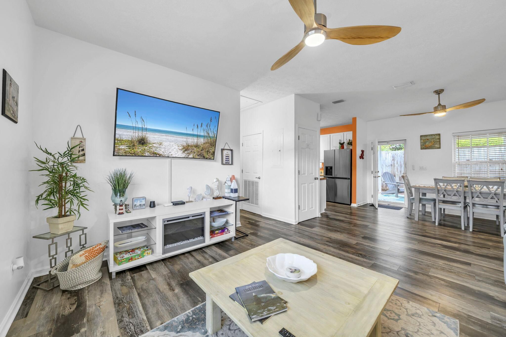 A spacious living area with a wooden floor, a ceiling fan, a dining table, and a kitchen in the background.