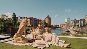 A group of people relaxing on a blanket by a riverside with historic buildings and a tower in the background.