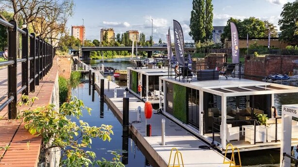 A floating restaurant with outdoor seating, a dock, and a view of the river and cityscape.