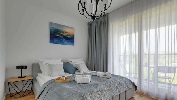 A modern bedroom with a large bed, a bedside table, a lamp, a painting on the wall, and sheer curtains. - Porto 42 | 2 Bedrooms | Gym | Balcony | Parking (Gdańsk)