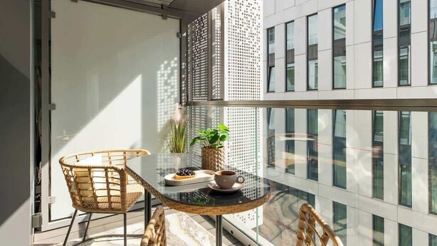 En balkong med ett glasbord, flätade stolar, en krukväxt och utsikt över moderna byggnader.