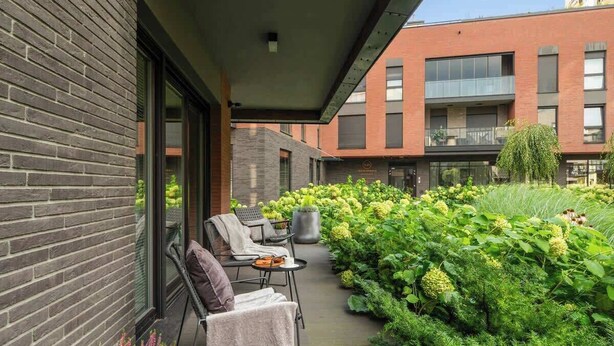 Ein modernes Gebäude mit Backsteinfassade, einem überdachten Außenbereich mit Stühlen und Tischen sowie einem üppigen Garten mit verschiedenen Pflanzen und einem kleinen Wasserspiel.