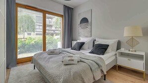Ein modernes Schlafzimmer mit einem großen Bett, einem Nachttisch mit Lampe und Blick auf Grünfläche.