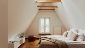 Ein Schlafzimmer mit schrägen Wänden, einem Bett mit weißer Bettwäsche, einem Fenster und einem kleinen Nachttisch mit Lampe.
