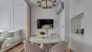 A modern living room with a white sofa, a round dining table with chairs, a TV mounted on the wall, and a chandelier.