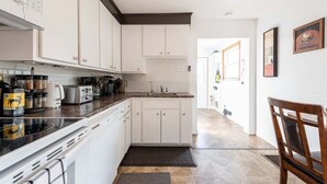 A modern kitchen with white cabinets, a black countertop, and a dishwasher. There is a dining area with a wooden chair and a small table.