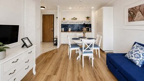 Uma sala de estar moderna com um sofá azul, uma mesa de jantar com cadeiras, uma área de cozinha e uma TV sobre um armário branco.