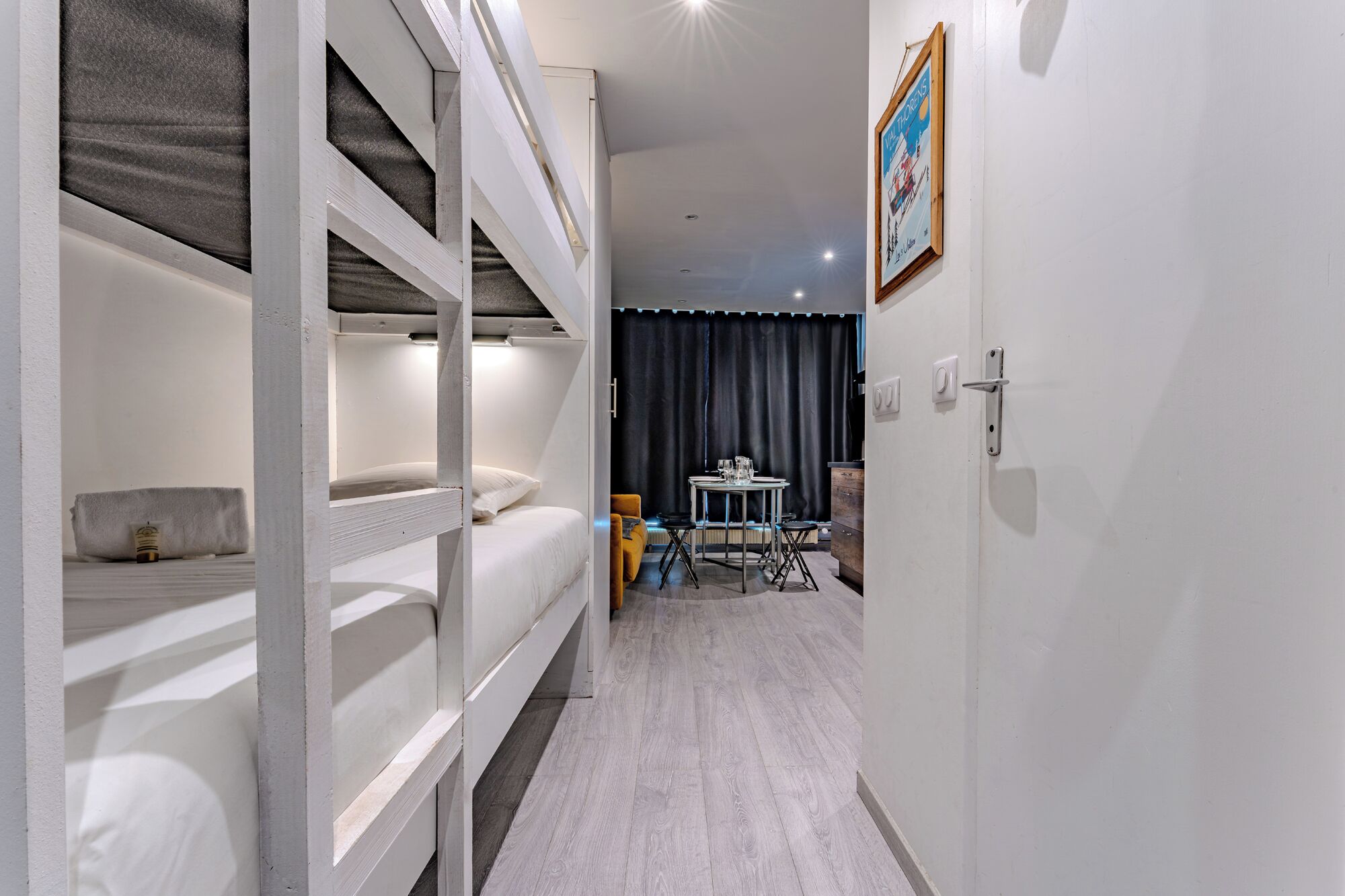 Habitación de hotel con literas, una mesita con sillas, una cortina oscura y un cuadro en la pared.
