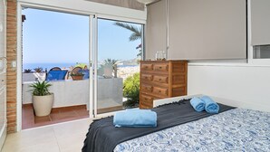 Un dormitorio con una cama, un aparador de madera, un ventanal corredero que da a un balcón y vistas al mar.