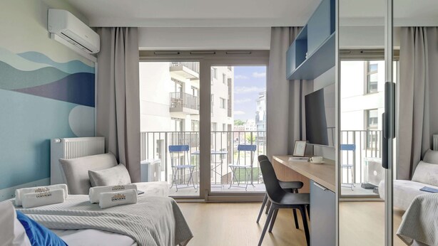 A hotel room with a bed, a desk, a chair, a TV, and a balcony with a view of other buildings. - Żurawie A31 | Shipyard | Foodhall Montownia (Gdańsk)