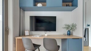 A modern workspace with a large flat-screen TV, two grey chairs, a wooden desk, and a blue shelving unit above. - Żurawie A31 | Shipyard | Foodhall Montownia (Gdańsk)