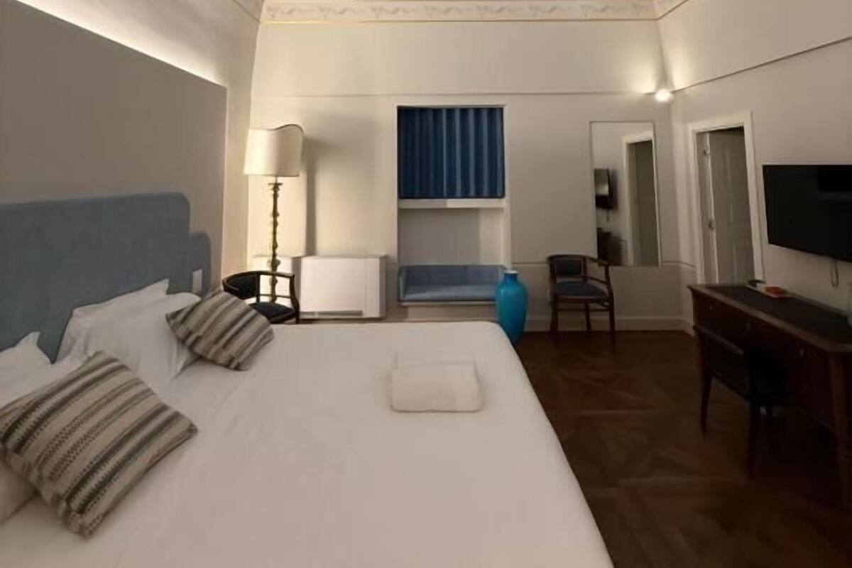 A hotel room with a large bed, a desk, a chair, a TV, and a small seating area.