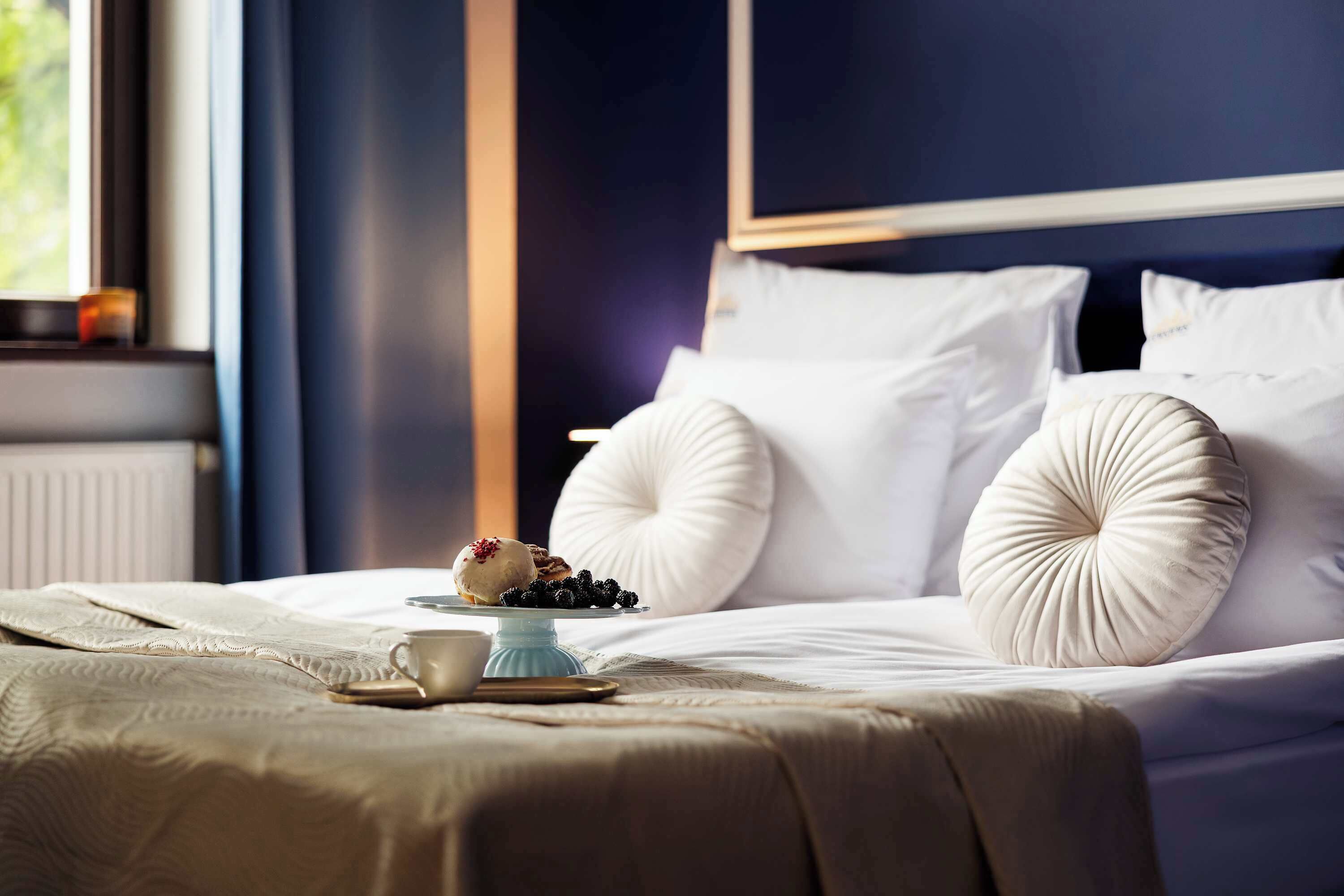 A neatly made bed with white linens, a small table with a cup and a plate of berries, and a window with a view of greenery.