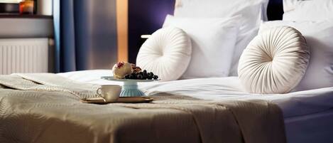 A neatly made bed with white linens, a small table with a cup and a plate of berries, and a window with a view of greenery.