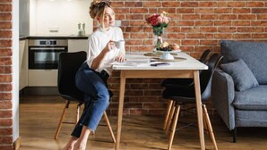Une personne est assise à une table à manger en bois, avec un vase de fleurs, dans une pièce dont le mur est en briques et qui contient un canapé gris.