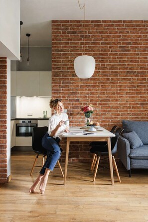 A person seated at a wooden dining table with a vase of flowers, in a room with a brick wall and a grey sofa. - Krowoderska 2 | Near Old Town (Kraków)