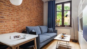 Une pièce chaleureuse avec un mur en briques, un canapé gris, une table blanche avec une tasse et une soucoupe, une petite table avec de la nourriture, et une fenêtre donnant sur un paysage verdoyant.