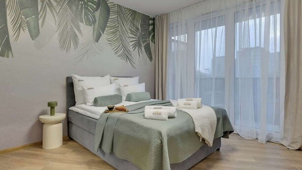 Een slaapkamer met een bed, nachtkastje, raam met gordijnen en een muur met bladmotief.