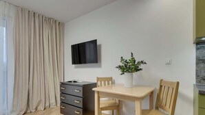 Een kleine kamer met een houten tafel, twee stoelen, een aan de muur bevestigde televisie, een donker kastje en een plant in een pot.