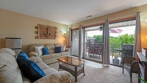 En stue med en sofa, et glasbord og en skydedør, der fører ud til en udendørs patio med en grill.