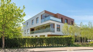 Ein modernes, mehrgeschossiges Wohngebäude mit Balkonen, blauem Himmel und einer gepflegten Hecke.