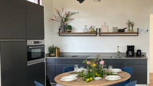 Una cocina moderna con armarios oscuros, horno empotrado, una mesa de comedor con sillas azules y varios objetos decorativos en estantes.