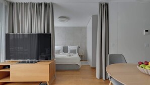 Une chambre d’hôtel avec un lit, une télévision, une commode en bois, une table à manger avec des fruits et des rideaux.