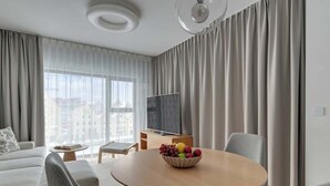 Un salon moderne avec une table ronde en bois, un canapé, un téléviseur et une grande fenêtre avec des rideaux.