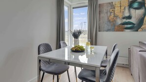 A modern dining area with a glass table, grey upholstered chairs, a vase with black stems, a bowl of fruit, and a glass of water. - Przystań Letnica 39 | Comfortable Stay & Parking (Gdańsk)
