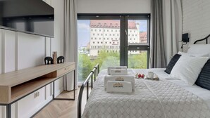 A modern hotel room with a large window, a bed with a quilted bedspread, a bedside table, a TV mounted on the wall, and a view of the cityscape.