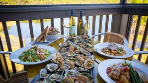 A dining table with various dishes, a bottle of wine, and a glass of white wine on a balcony overlooking a body of water. - Crisp Ocean Breeze | Private Pool | Walk to Beach | Golf Cart(LSV) (Santa Rosa Beach)
