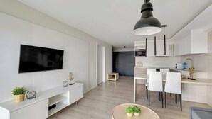 Uma sala de estar moderna com televisão de tela plana, área de jantar com cadeiras brancas, cozinha com armários brancos e uma luminária pendente grande.