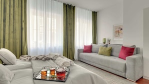 Ein modernes Hotelzimmer mit einem Bett, einem grauen Sofa, einem Couchtisch mit Tablett und grünen Vorhängen.